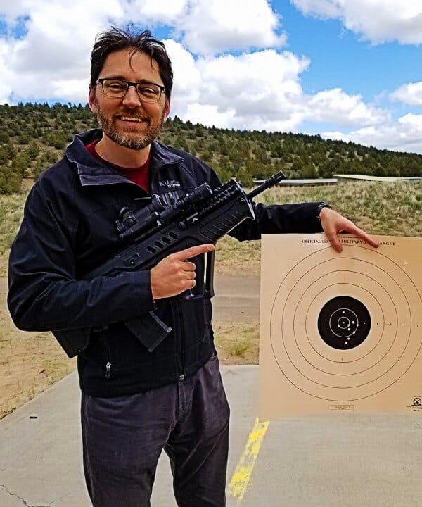Oregon self-defense attorney Michael Romano with a scoped bullpup rifle beside a target showing a tight shot group