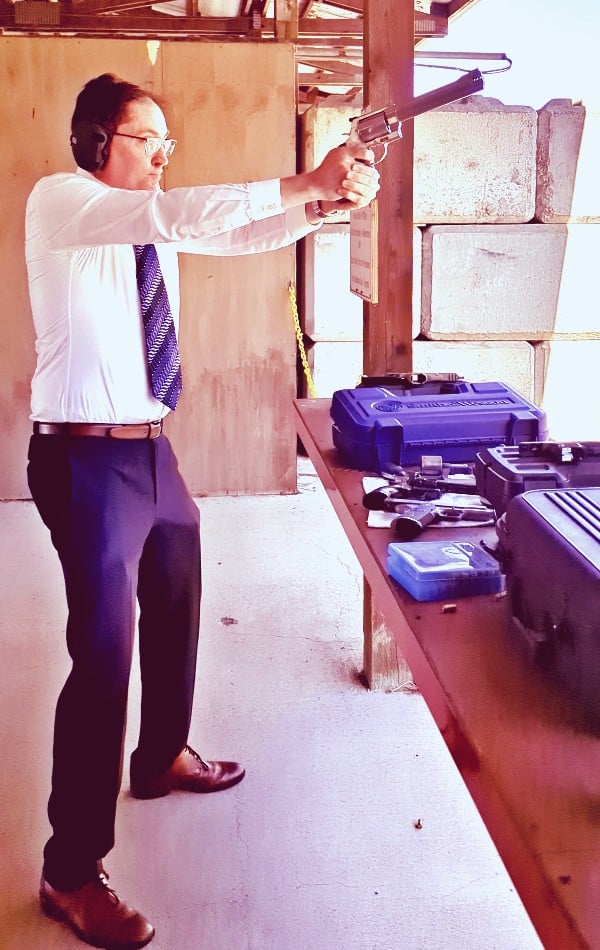 Oregon gun rights attorney Michael Romano firing a Smith & Wesson 460XVR revolver at a shooting range beside locked hard-sided pistol cases.
